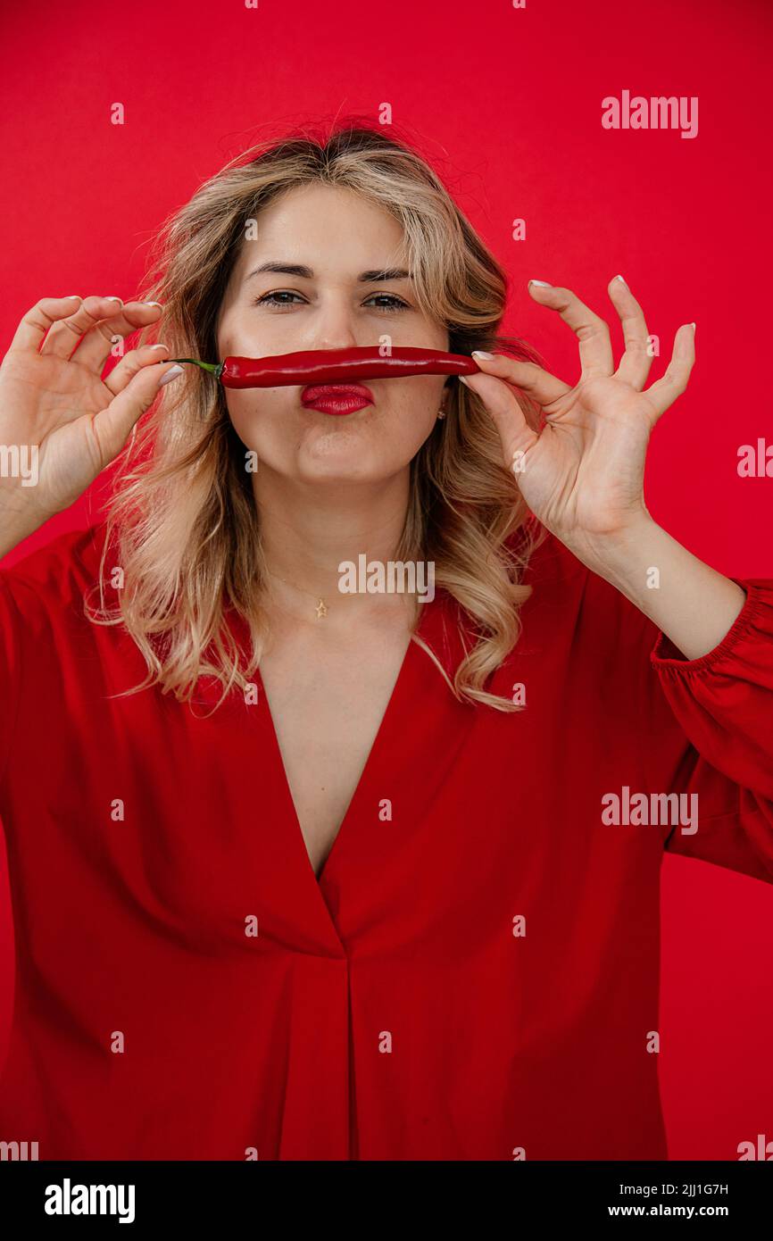 Vertical funny, creative blonde woman in red outfit holding red hot pepper in hands under nose like mustache. Hot news Stock Photo