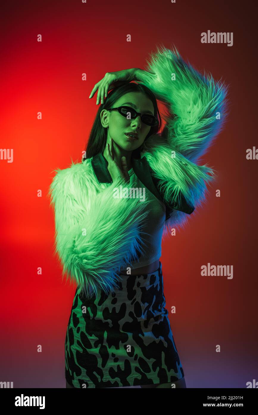 stylish young woman in faux fur jacket and sunglasses posing on red ...
