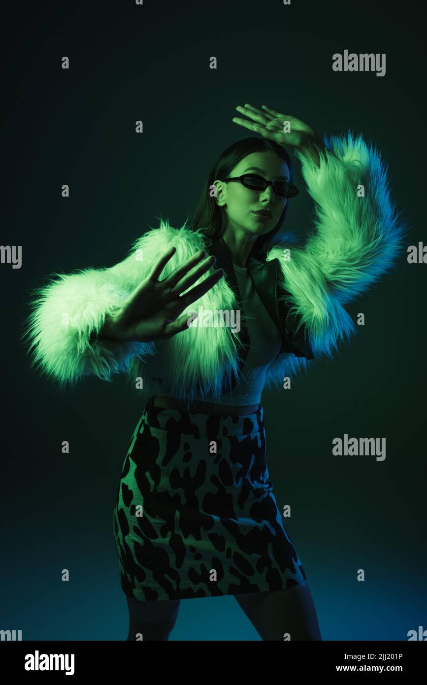 stylish young woman in faux fur jacket and sunglasses gesturing while ...