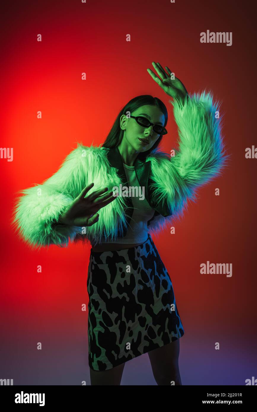 stylish young woman in faux fur jacket and sunglasses gesturing while ...
