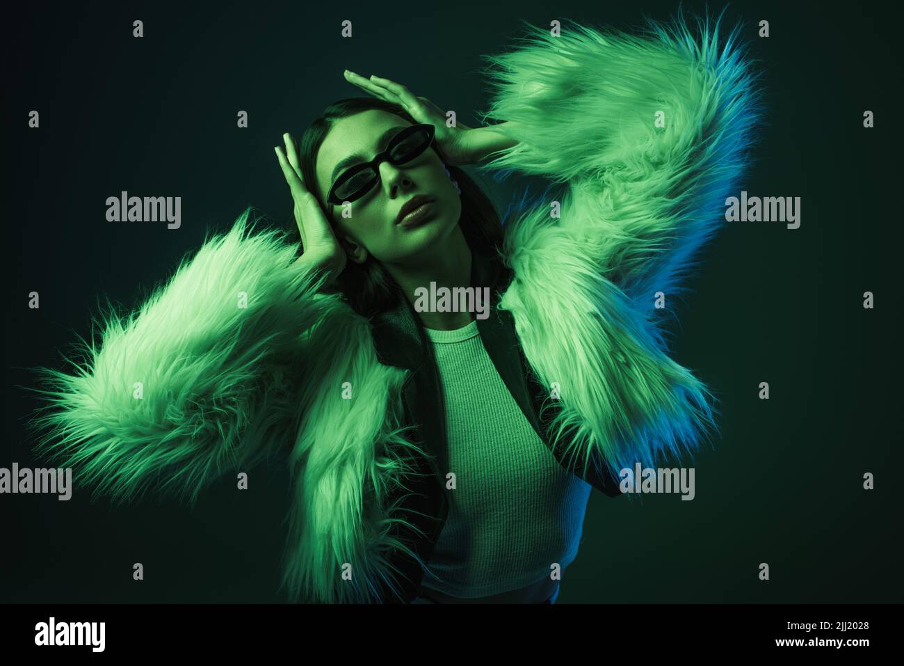 stylish young woman in faux fur jacket and sunglasses posing on blue ...