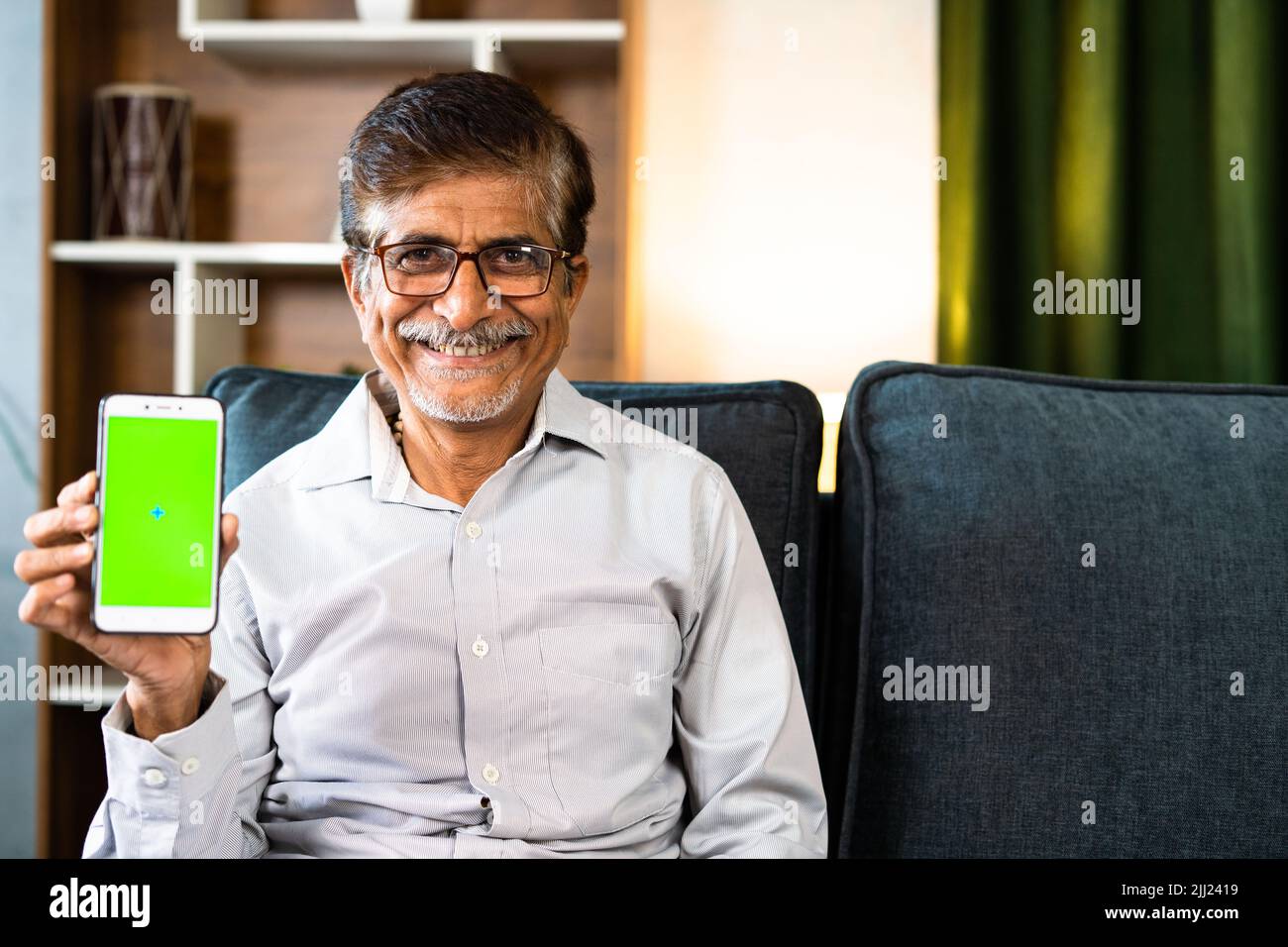 Happy smiling senior man showing green screen mobile phone while sitting on sofa at home with copy space - concept of online app or service promotion Stock Photo