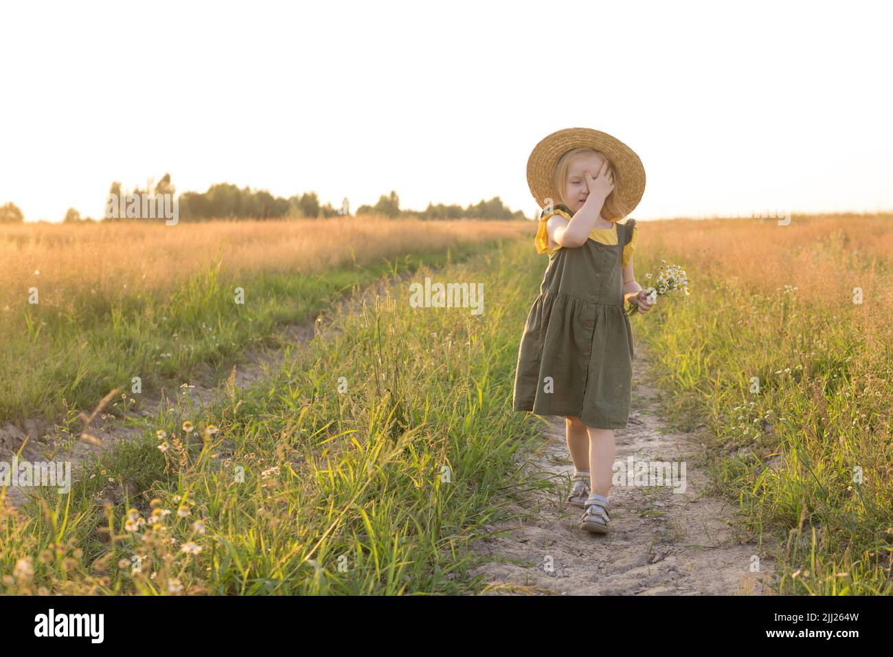 A little blonde girl in a straw hat walks in a field with a bouquet of ...