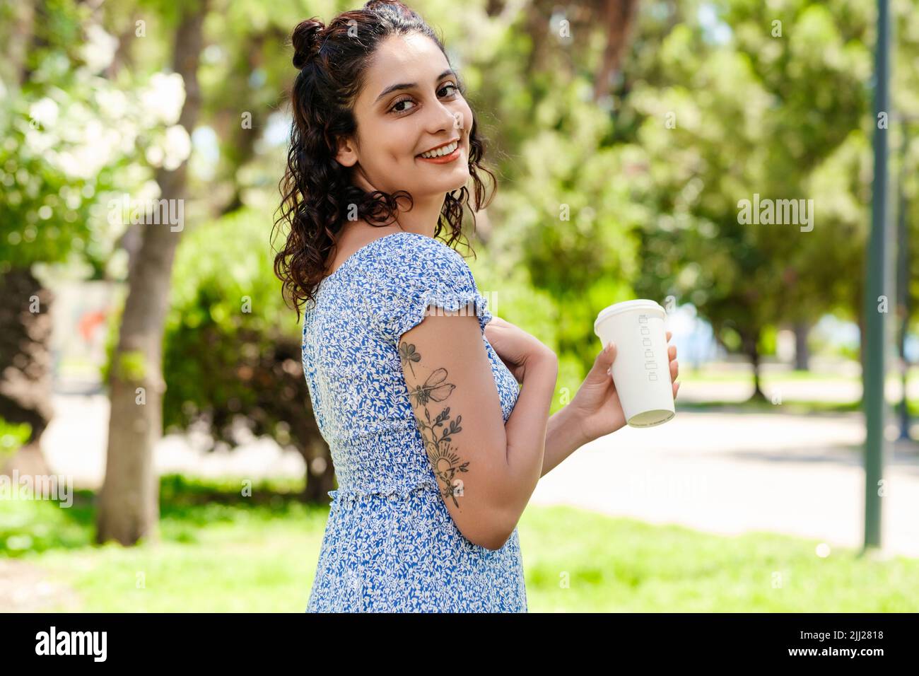 Side view of brunette woman wearing summer dress standing on city park, outdoors posing and looking at the camera while holding a takeaway cup of coff Stock Photo