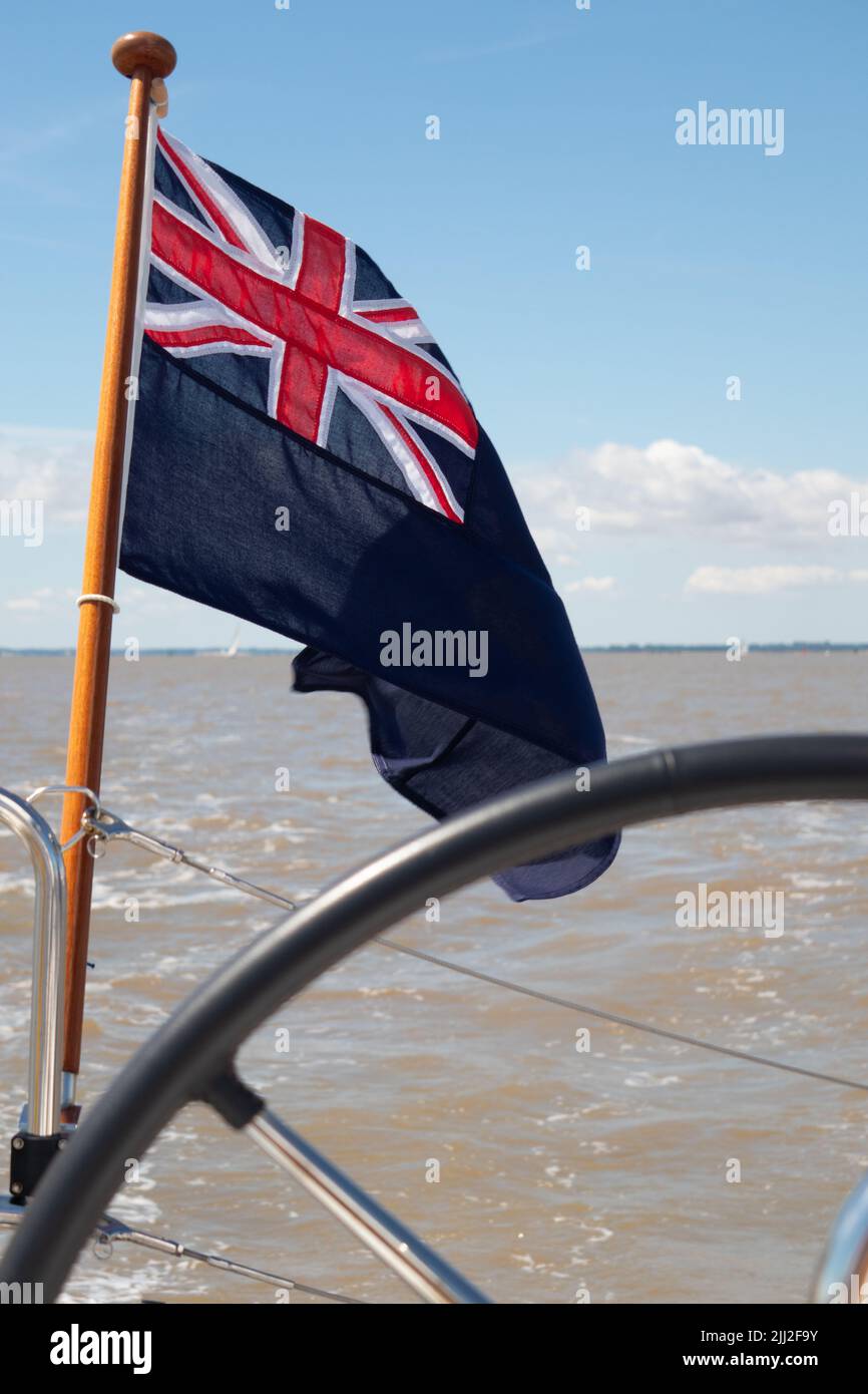 Blue Ensign on a wooden flagstaff flying on a stern of a yacht showing part of the wheel and sea in background Stock Photo