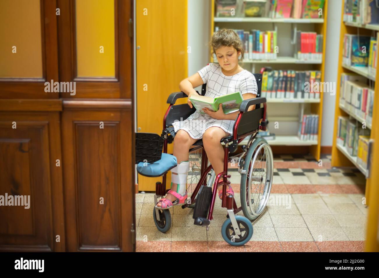 A little disabled girl in a wheelchair reads books with pleasure. Life of children with special needs. Happy education concept for disabled children Stock Photo