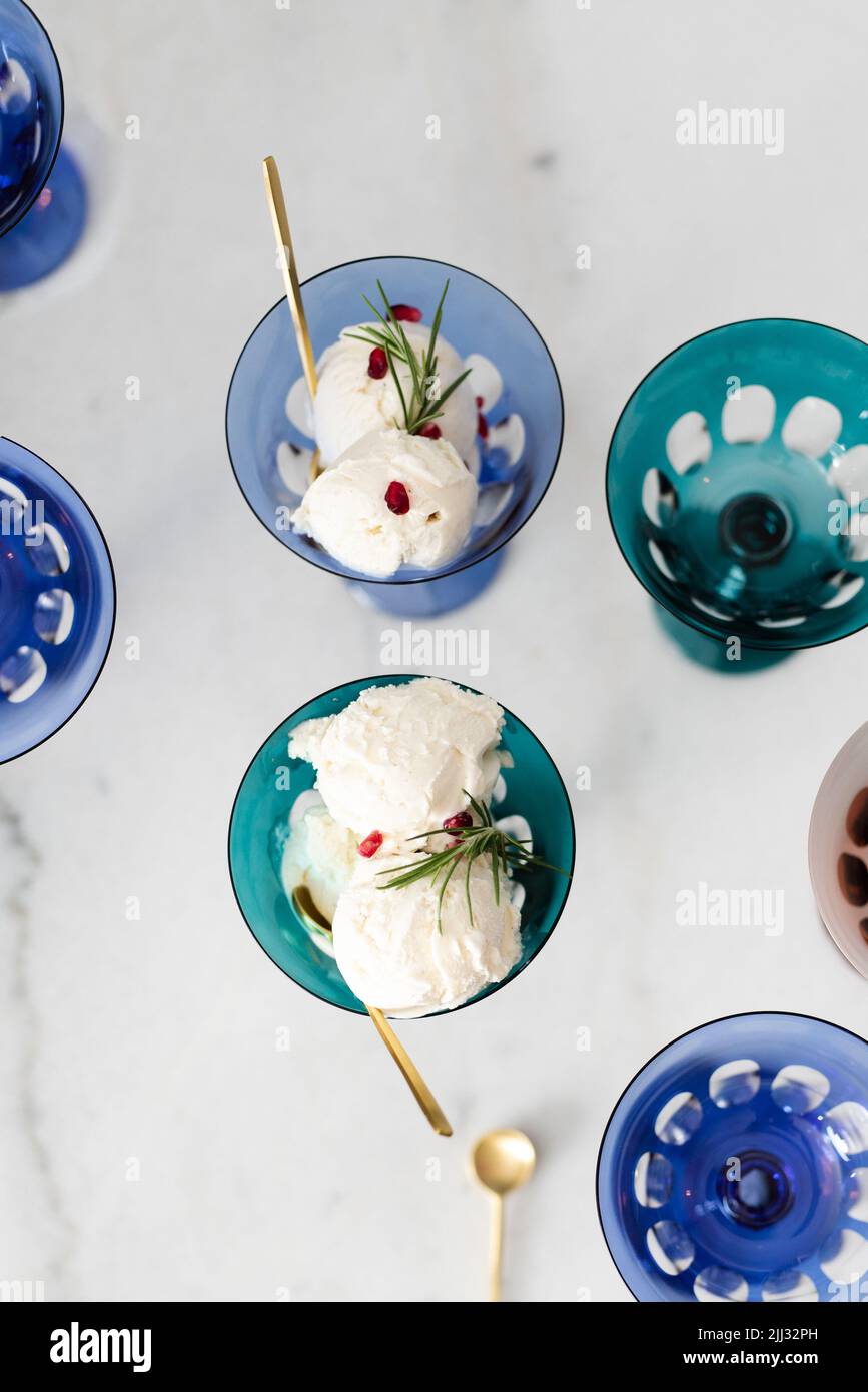 Vertical flatlay view of colorful clear blue and green wine glasses with ice cream and gold spoons against white marble. Stock Photo