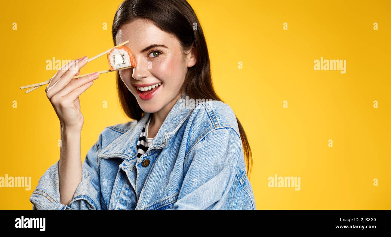 Beautiful modern girl posing with sushi roll and chopsticks on orange background Stock Photo