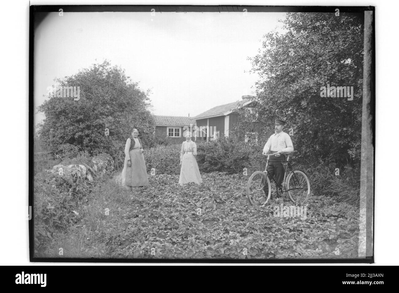 One -storey residential building and outbuildings, two women and a man with bicycle.Petrus Johansson Stock Photo