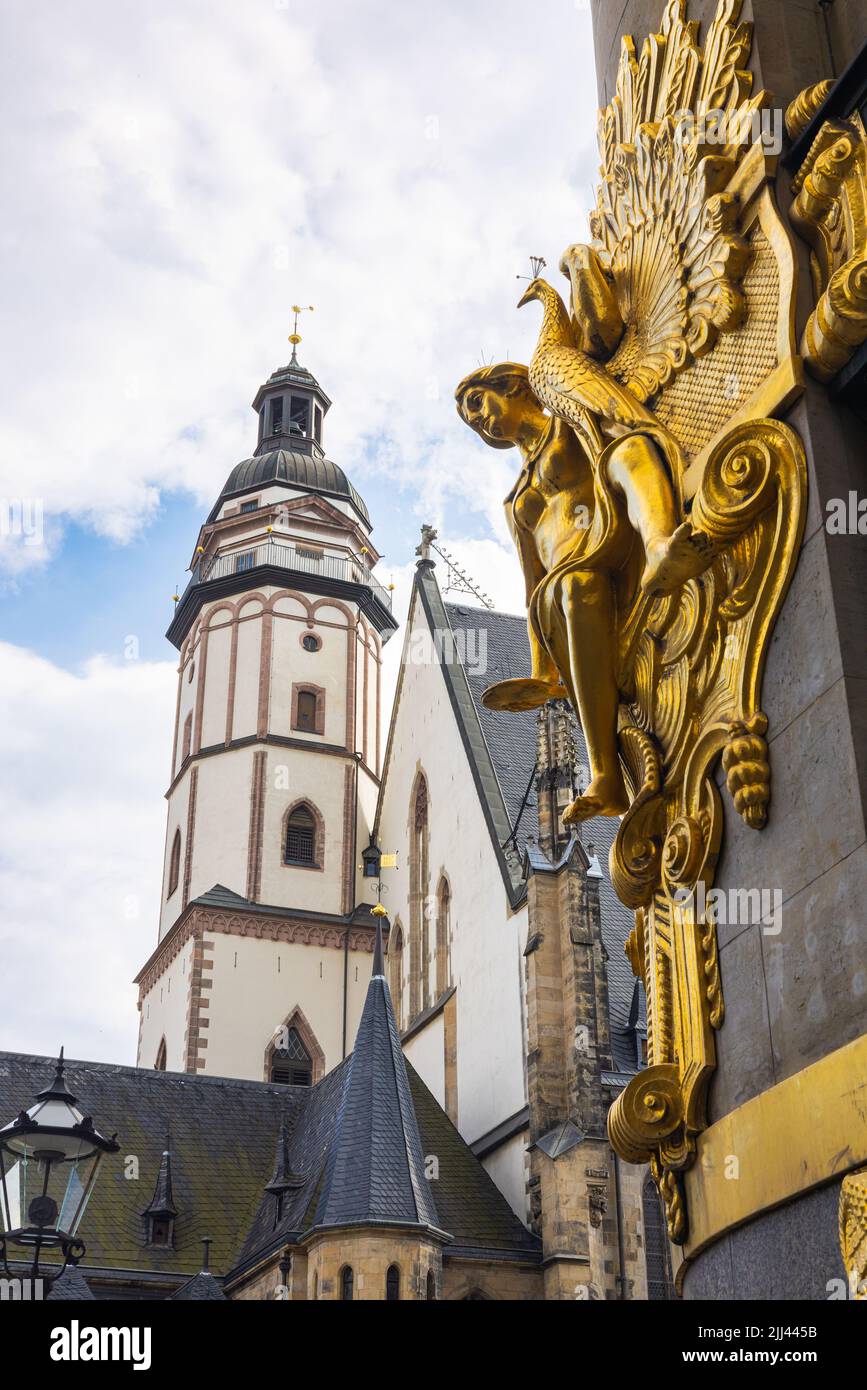 Leipzig, Germany - June 25, 2022: The the bell tower of the Thomaskirche. The golden sculpture of the Commerzbank or former Konfektionshaus Ebert blur Stock Photo