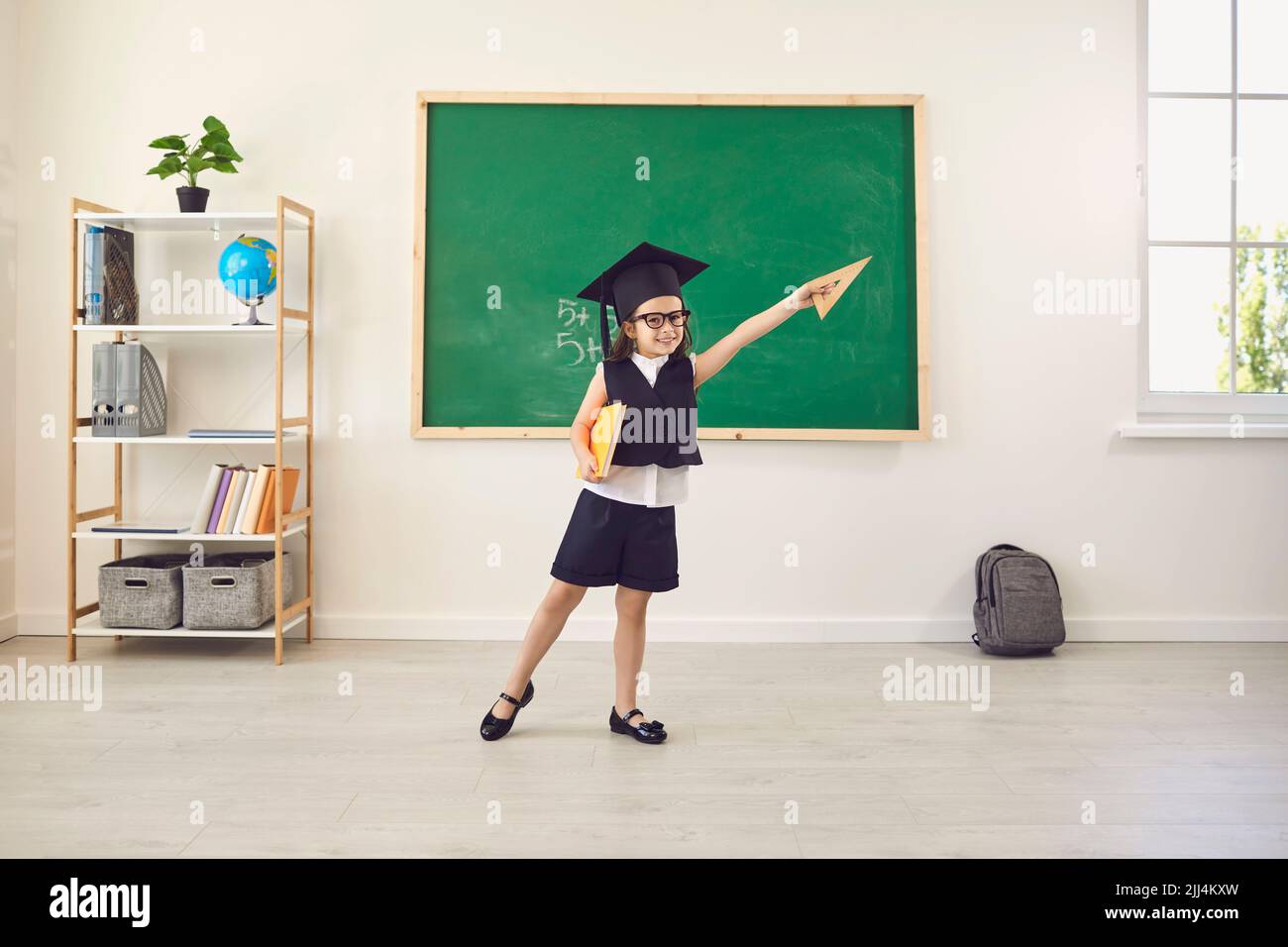 Back to school. Funny schoolgirl in a graduation hat raised her hand up in the classroom. Stock Photo