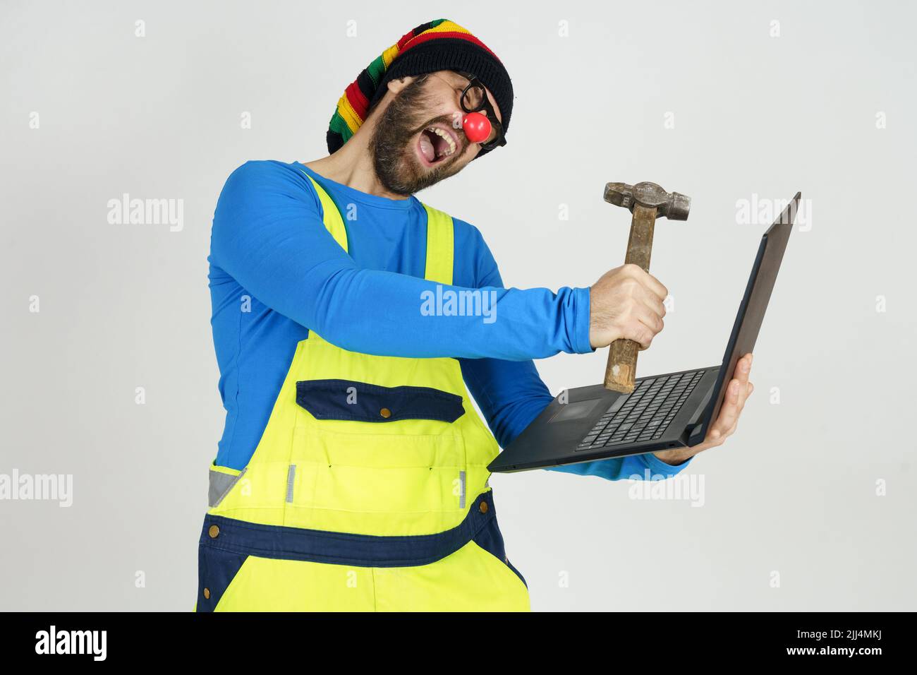 Humor and communication concept. A clown in a bright suit holds a laptop in his hands, hits it with a hammer Stock Photo