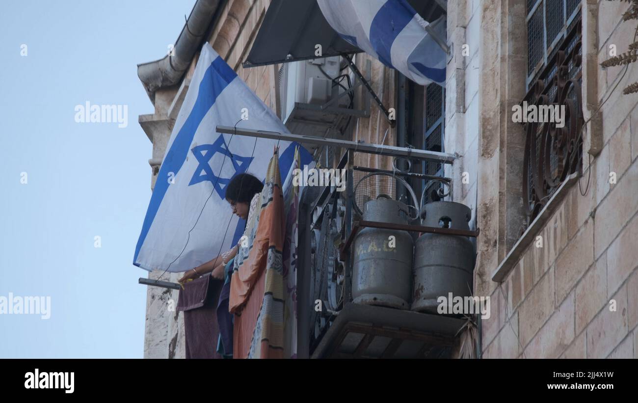 A Jewish settler hangs laundry on a balcony adorned with the Israeli flag in the Muristan area which is a complex of streets and shops in the Christian Quarter of the Old City on July 20, 2022 in Jerusalem, Israel. Previously, Greek Orthodox Patriarch of Jerusalem Patriarch Theophilos III told US President, Joe Biden, that there is an effort by Israeli right wing organizations to drive Christians out of the old city of Jerusalem, through property seizures. About 300 Jewish settlers already live in the Christian Quarter. Stock Photo