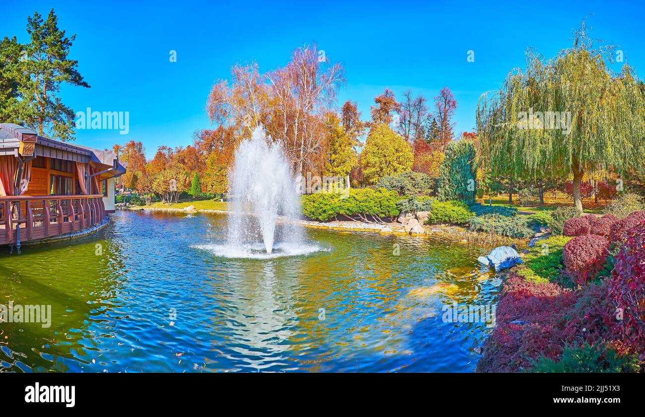 Panorama of the lake with beautiful fountain and lush autumn park around it with red barberry bushes, green conifer shrubs and yellow birches and oaks Stock Photo