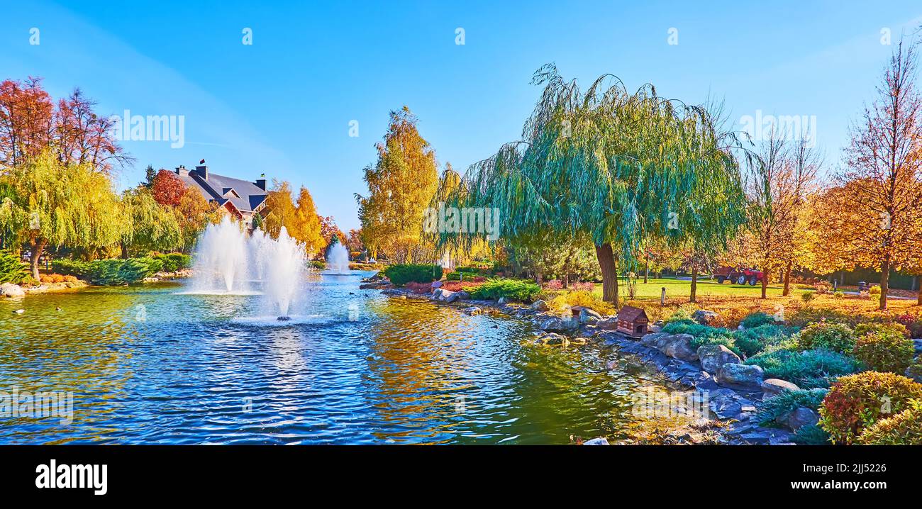 Panoramic view of the lake with fountains, junipers, willow trees, yellow autumn birches in background, Mezhyhirya, Ukraine Stock Photo