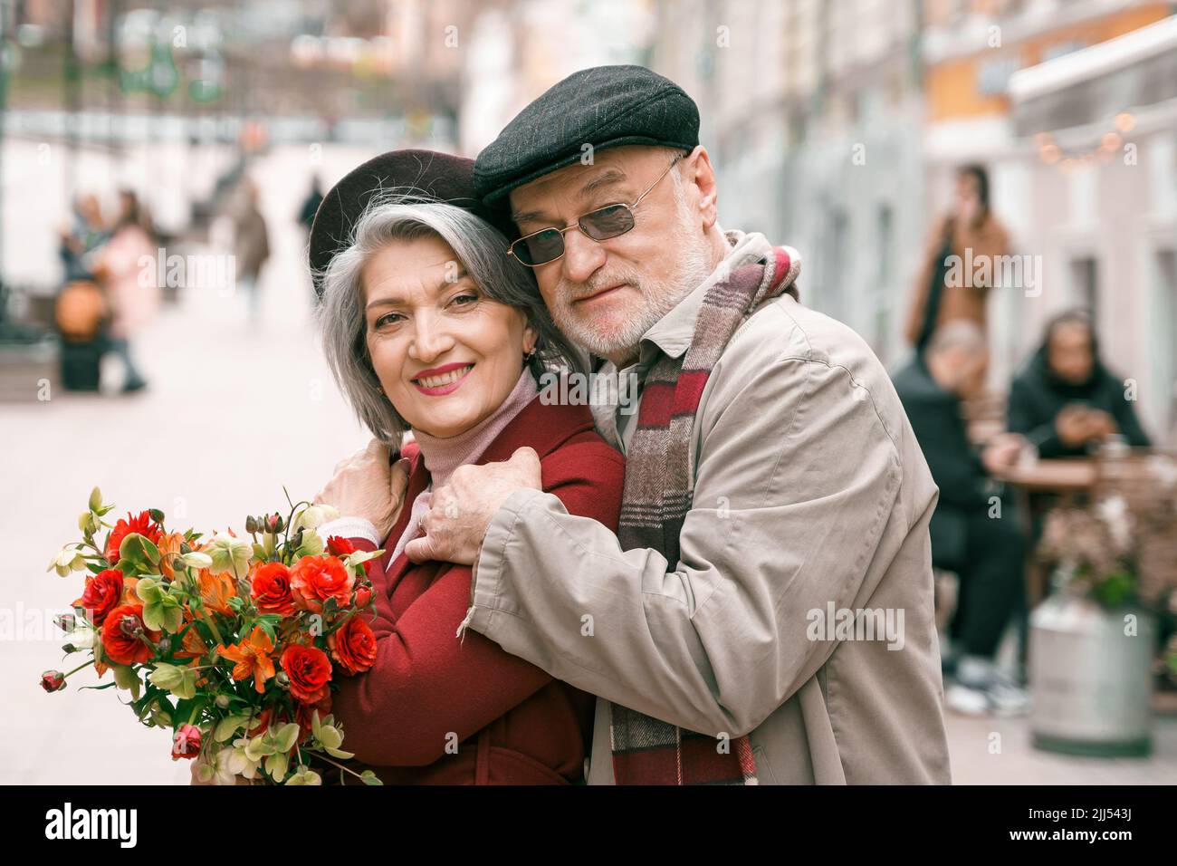 Elderly senior love couple. Old retired man woman together on romantic date.Aged husband wife walking on city street with flowers.Stylish elder huggin Stock Photo