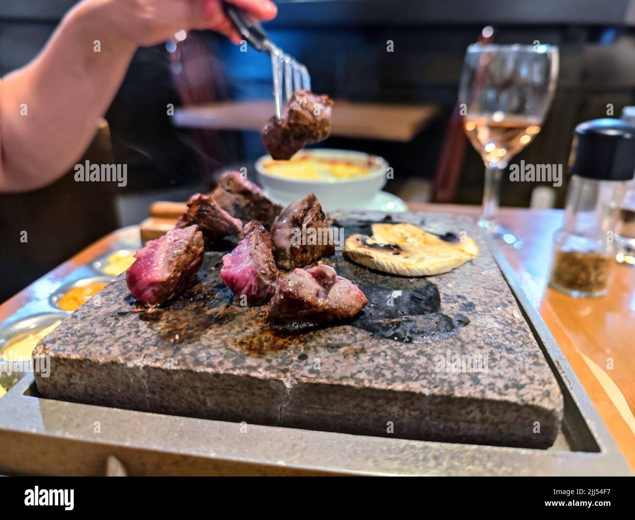 White woman's hand with a fork cooks steak on a stone grill in a restaurant on a dinner table Stock Photo