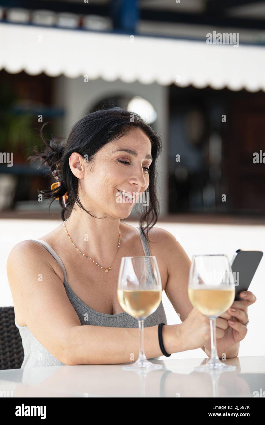 Adult woman sitting in an outdoor restaurant using her cell phone with two glasses of wine on the table Stock Photo