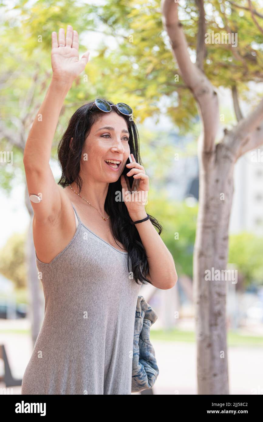 Adult woman with a glucose sensor device on her right arm talking with phone and greeting someone in a sunny day Stock Photo