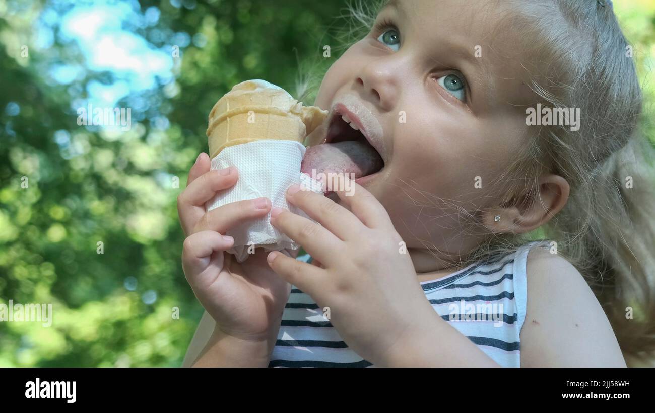 Cute little girl eats ice cream outside. Close-up portrait of blonde girl sitting on park bench and eating icecream. Stock Photo