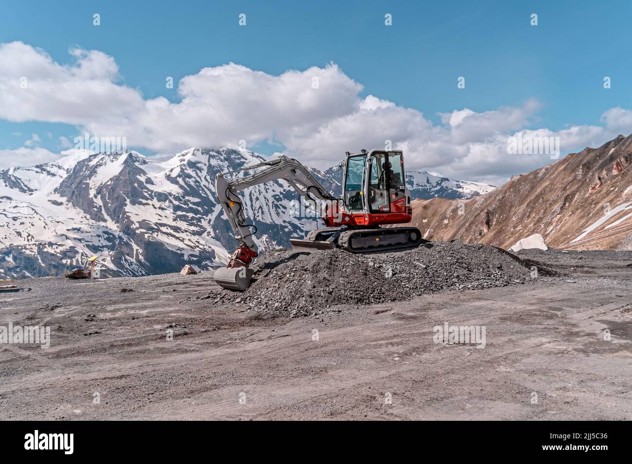 red excavator with shovel standing on hill with stones, mountains in background Stock Photo