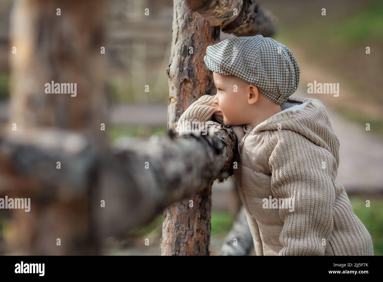 Bully boy kid with a slingshot aims at someone near a fence in the village outdoors. Rustic barefoot child boy with a hat shoots a slingshot. Stock Photo