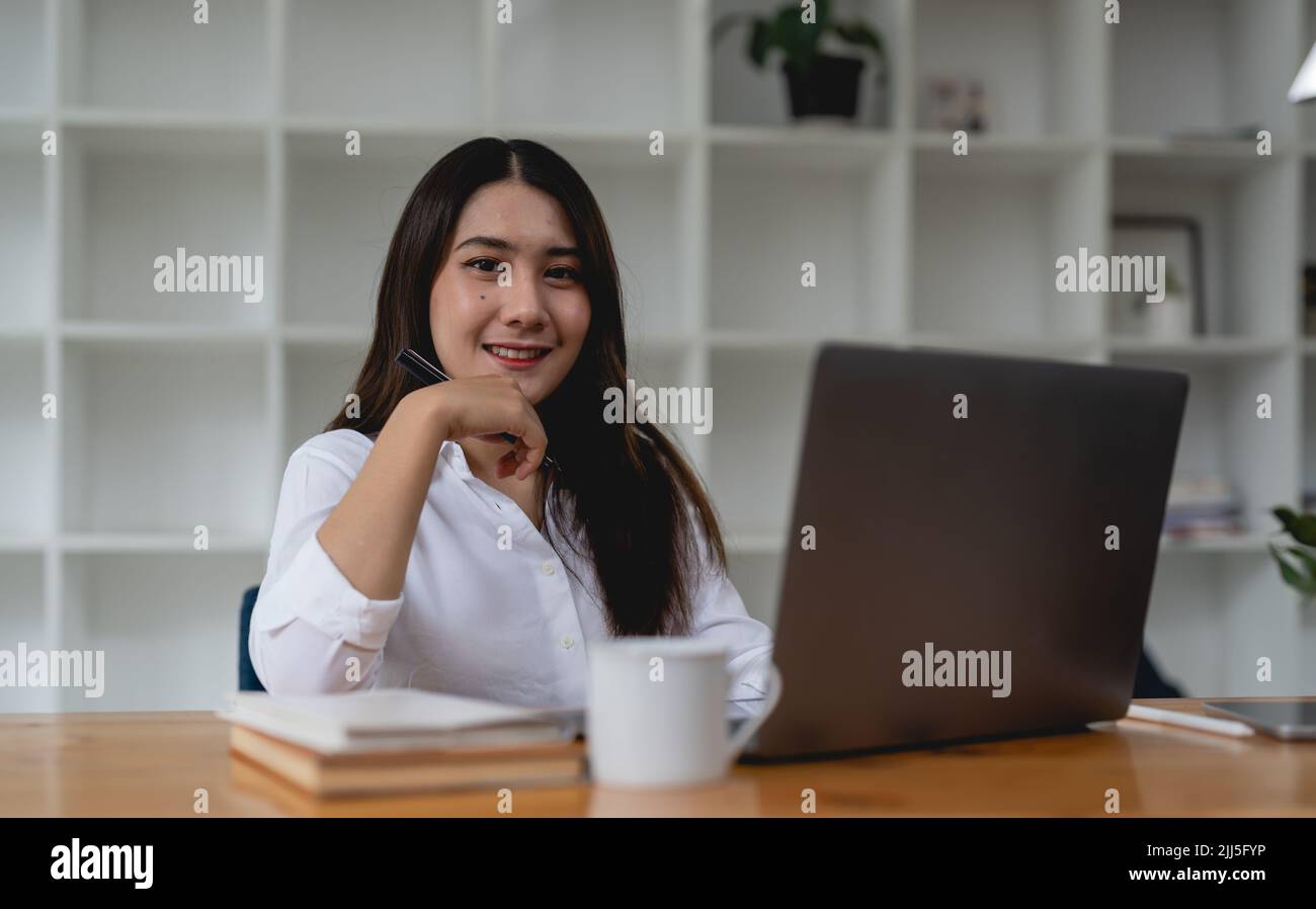 Image of joyful asian female worker wearing white shirt smiling while sitting at table in office, and working on laptop Stock Photo