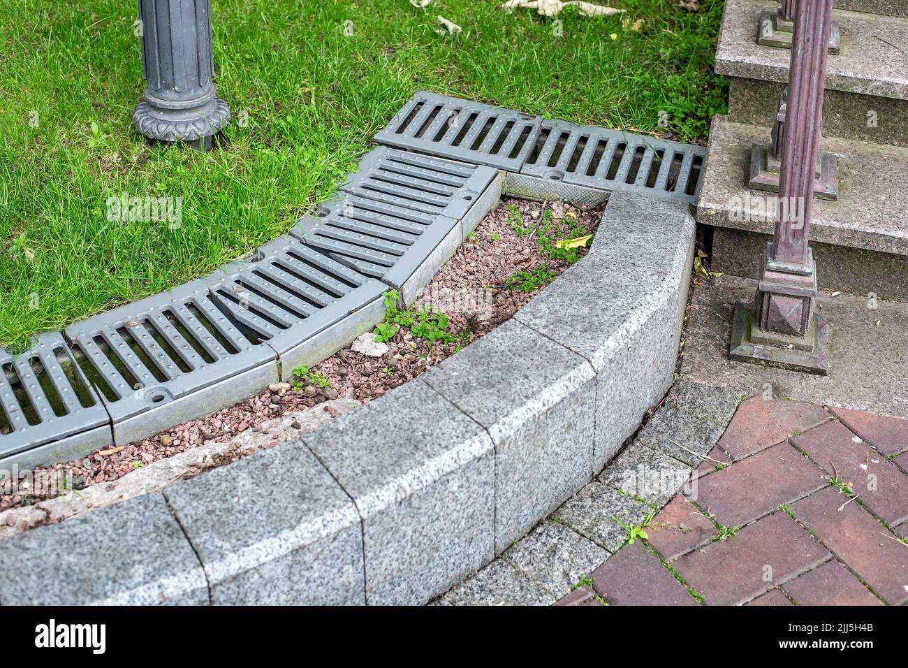 iron drainage grate on the roadside with green grass and stone pebbles at the granite curb along the pedestrian pavement of stone tiles near stairs wi Stock Photo
