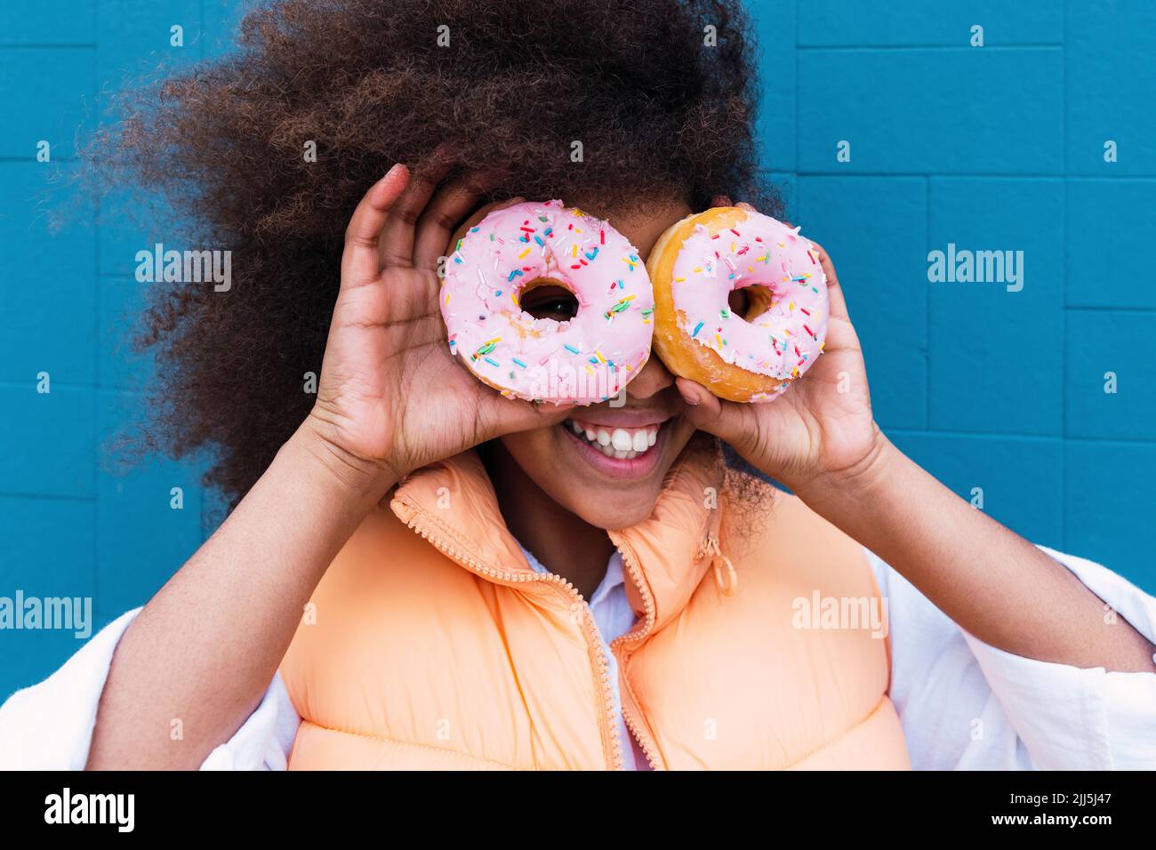Cheerful girl covering eyes with doughnuts in front of blue wall Stock Photo