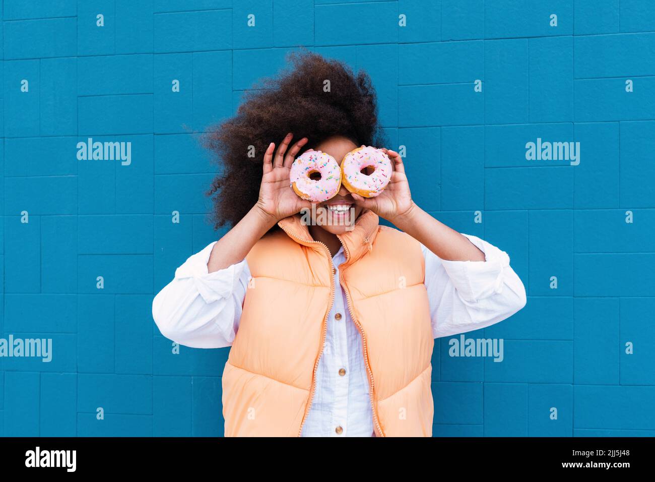 Happy girl covering eyes with doughnuts standing in front of blue wall Stock Photo