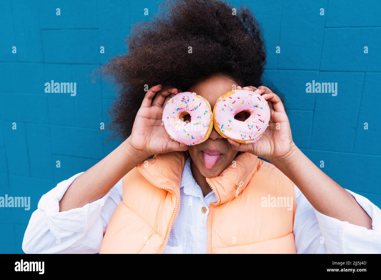 Girl sticking out tongue covering eyes with doughnuts in front of blue wall Stock Photo