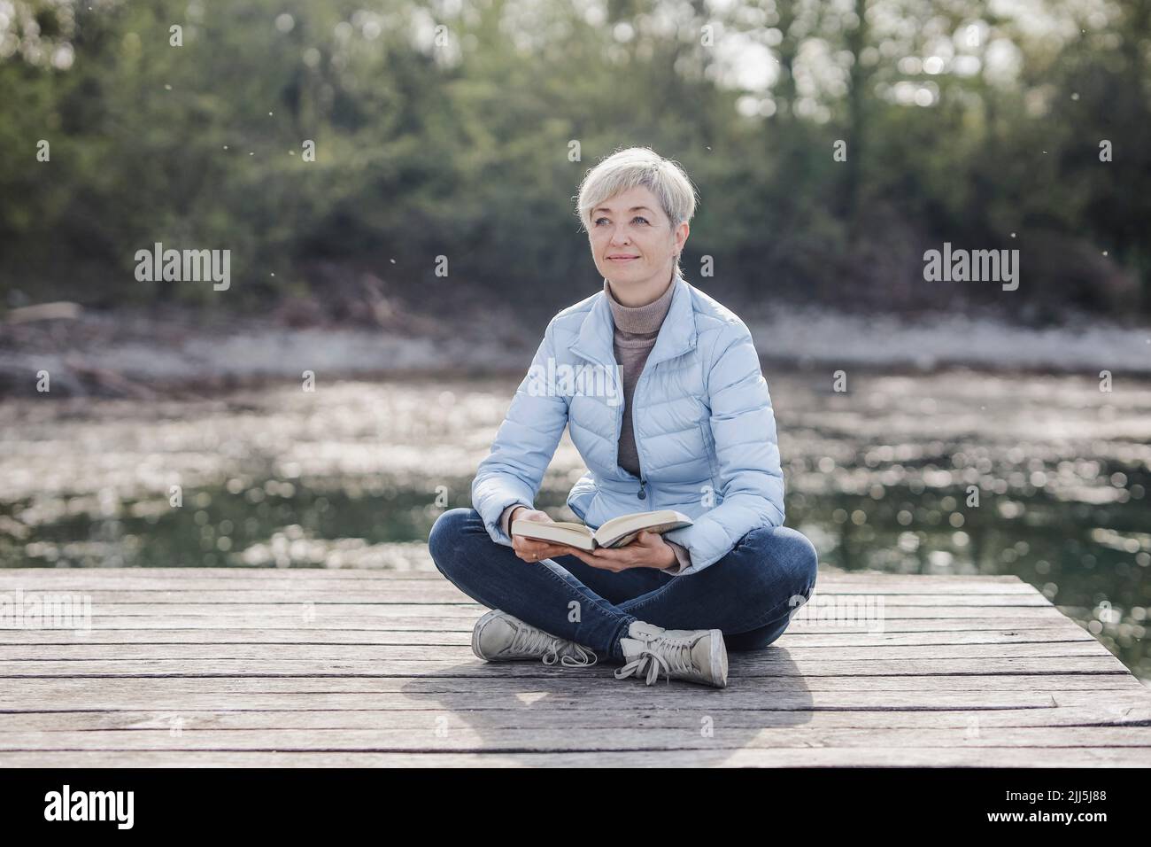 Mature woman sitting cross-legged with book on pier Stock Photo