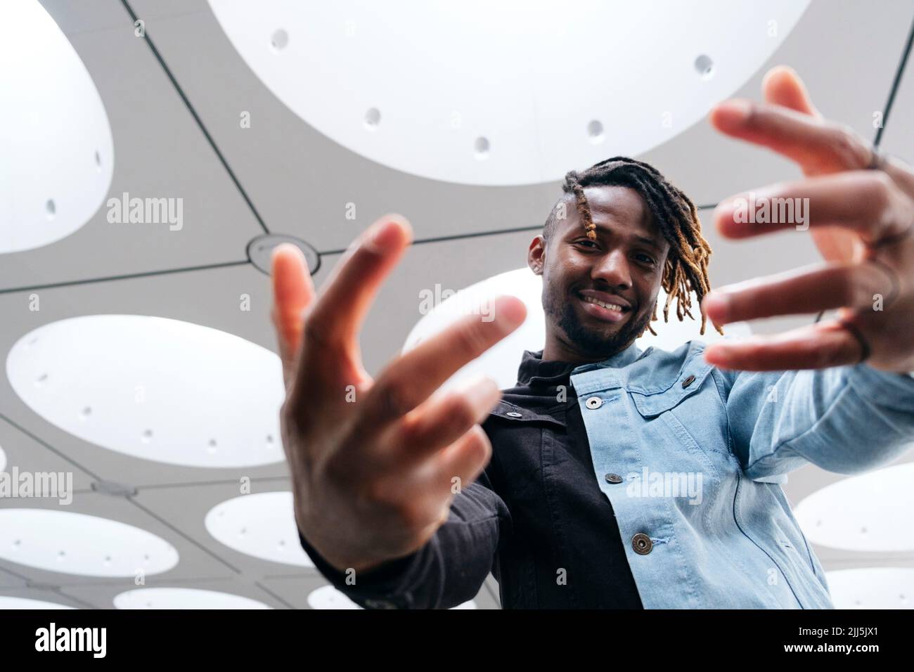 Smiling man with dreadlocks gesturing under illuminated ceiling Stock Photo