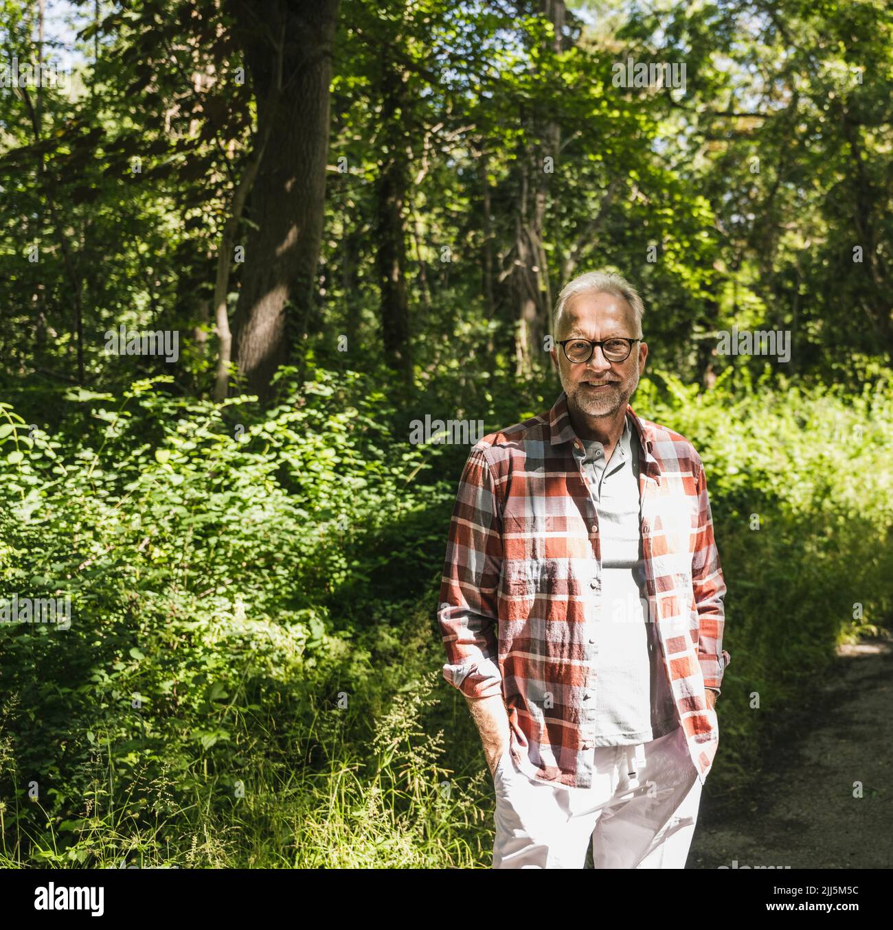 Smiling man with hands in pockets standing by plants on sunny day Stock Photo