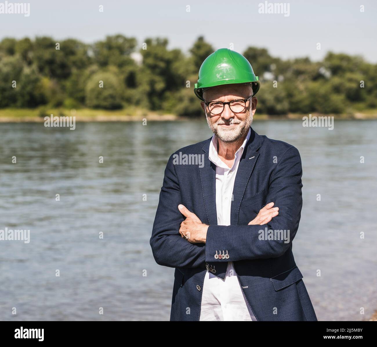 Smiling senior engineer wearing green hardhat standing with arms crossed at riverbank on sunny day Stock Photo