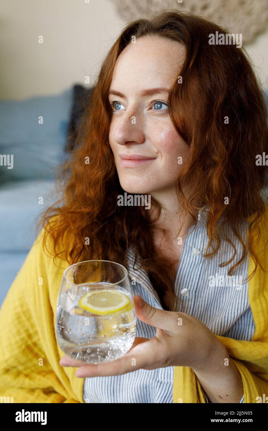 Contemplative woman holding glass of sparkling water with lemon slice Stock Photo