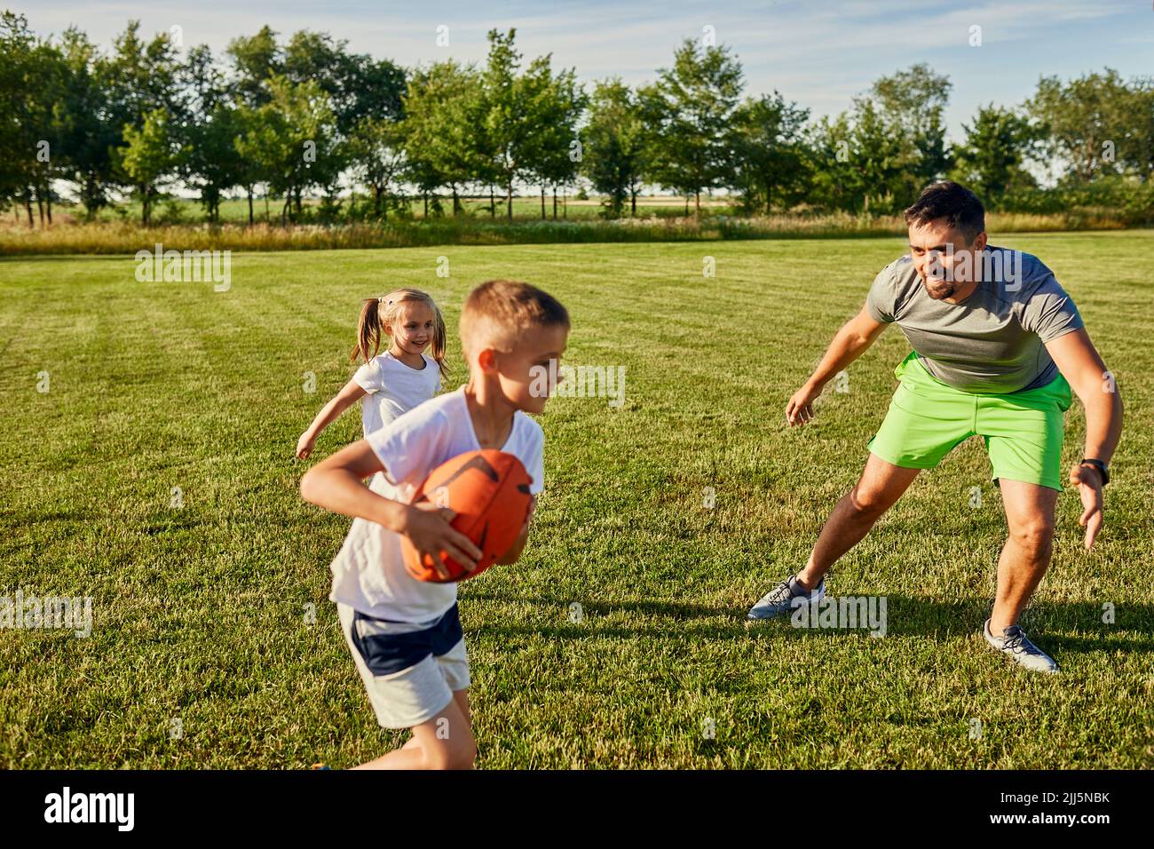 Father playing rugby with son and daughter at sports field on sunny day Stock Photo