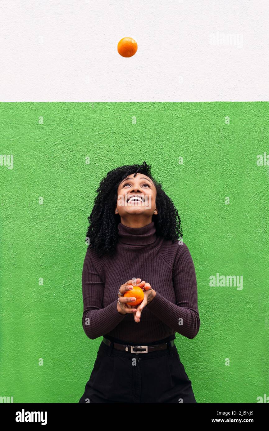 Happy woman throwing orange in front of green wall Stock Photo Alamy