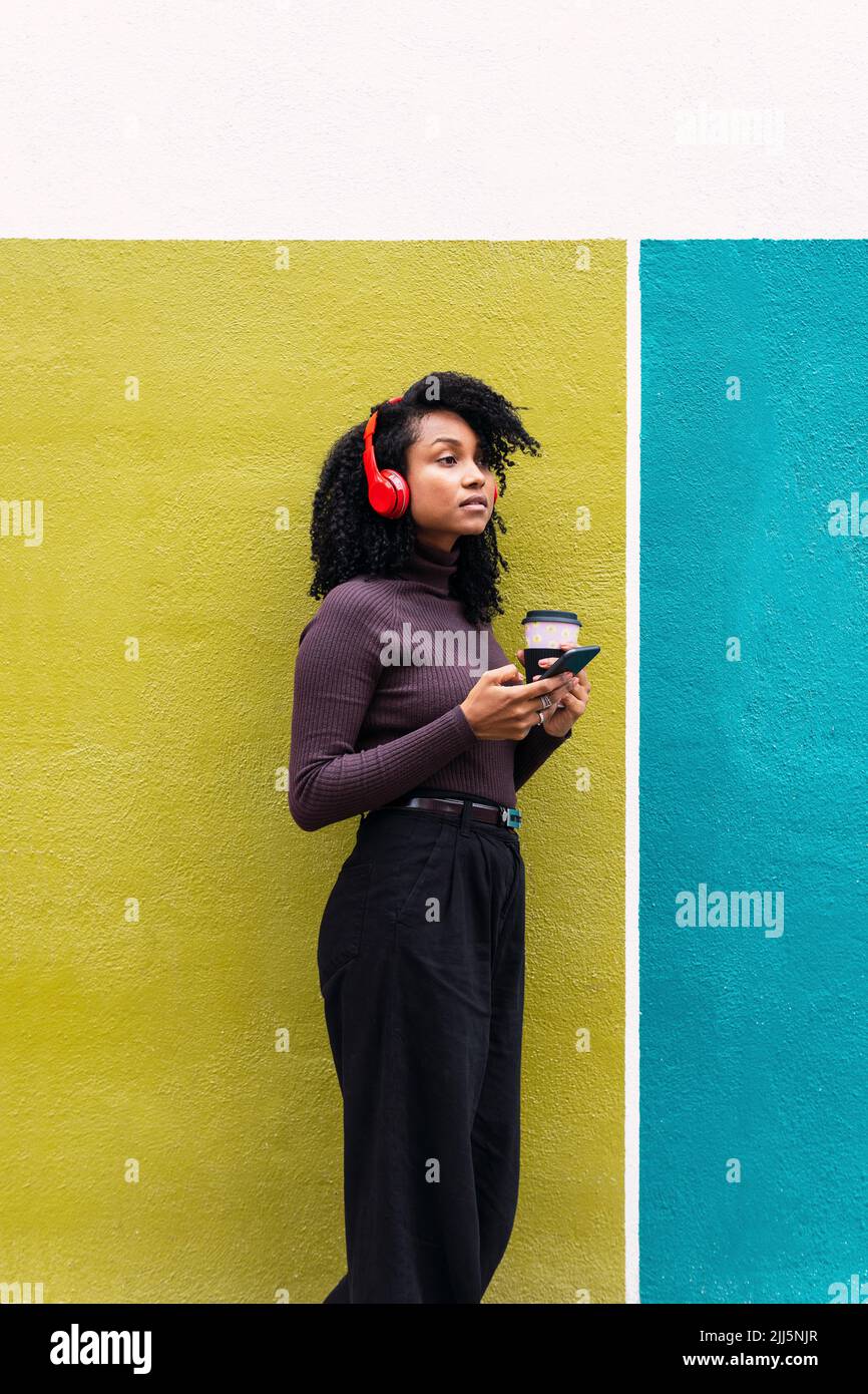 Woman wearing wireless headphones holding mobile phone and disposable coffee leaning on yellow and blue wall Stock Photo