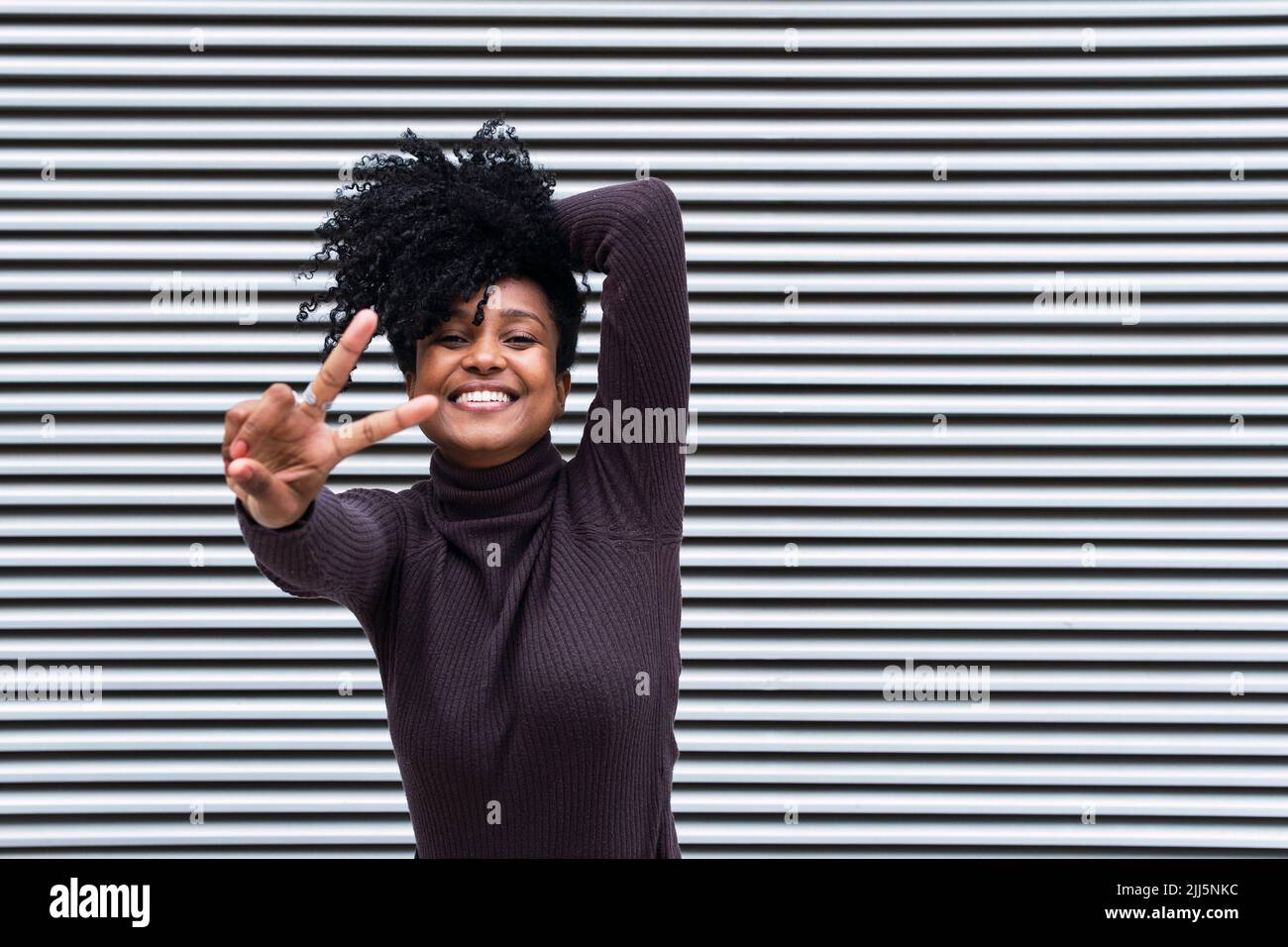 Happy woman with curly hair gesturing peace sign in front of wall Stock Photo