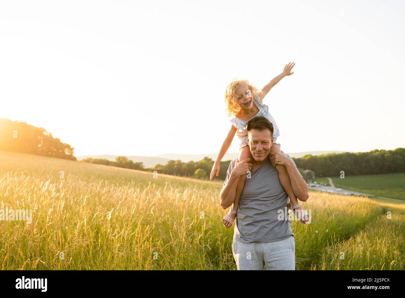 Smiling man carrying daughter sitting with arms outstretched at field Stock Photo