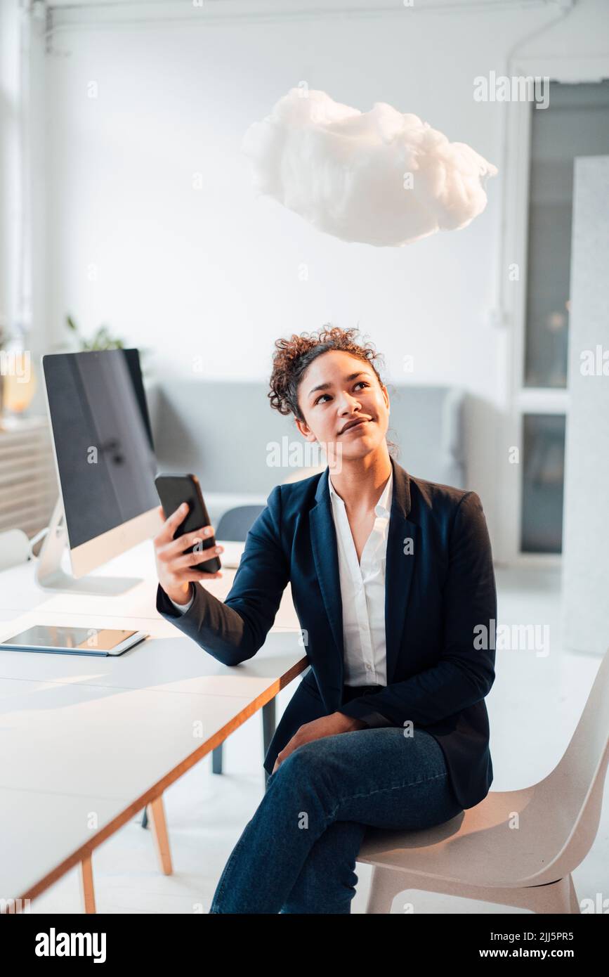 Smiling young businesswoman holding mobile phone looking at levitating cloud in office Stock Photo
