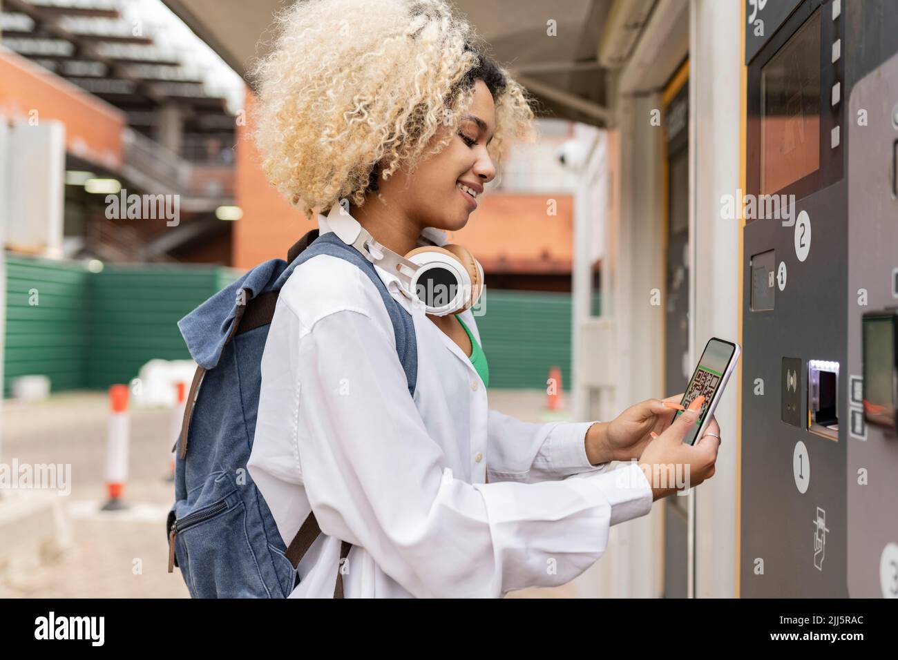 Smiling woman scanning QR code through smart phone on ticket vending machine Stock Photo