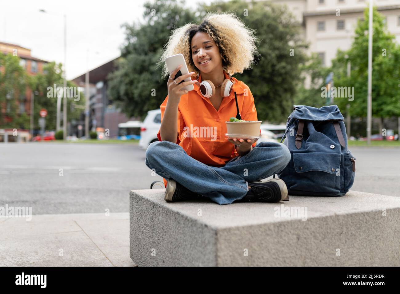 Smiling woman using smart phone holding lunch box sitting cross-legged on seat Stock Photo