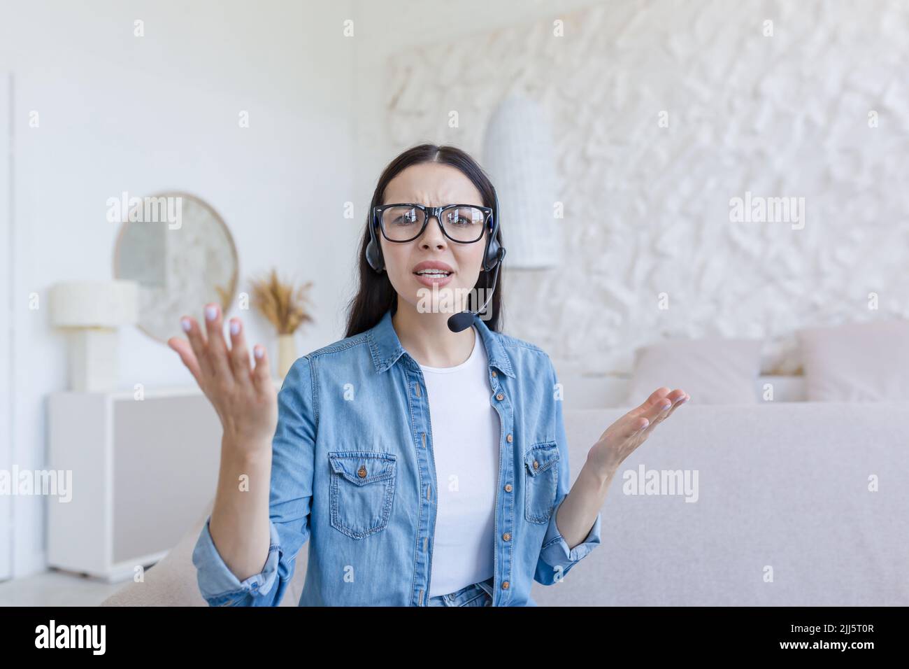 Beautiful woman looking at web camera, nervous and arguing with interlocutor, using headset for video call, working at home sitting on sofa, online conference and study Stock Photo
