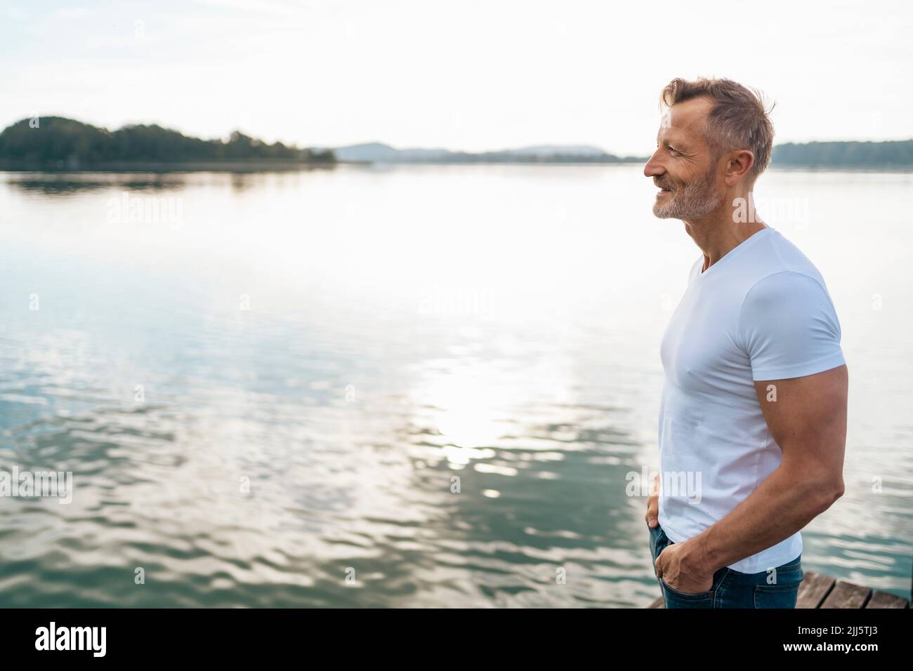 Smiling mature man with hands in pockets standing at lake Stock Photo