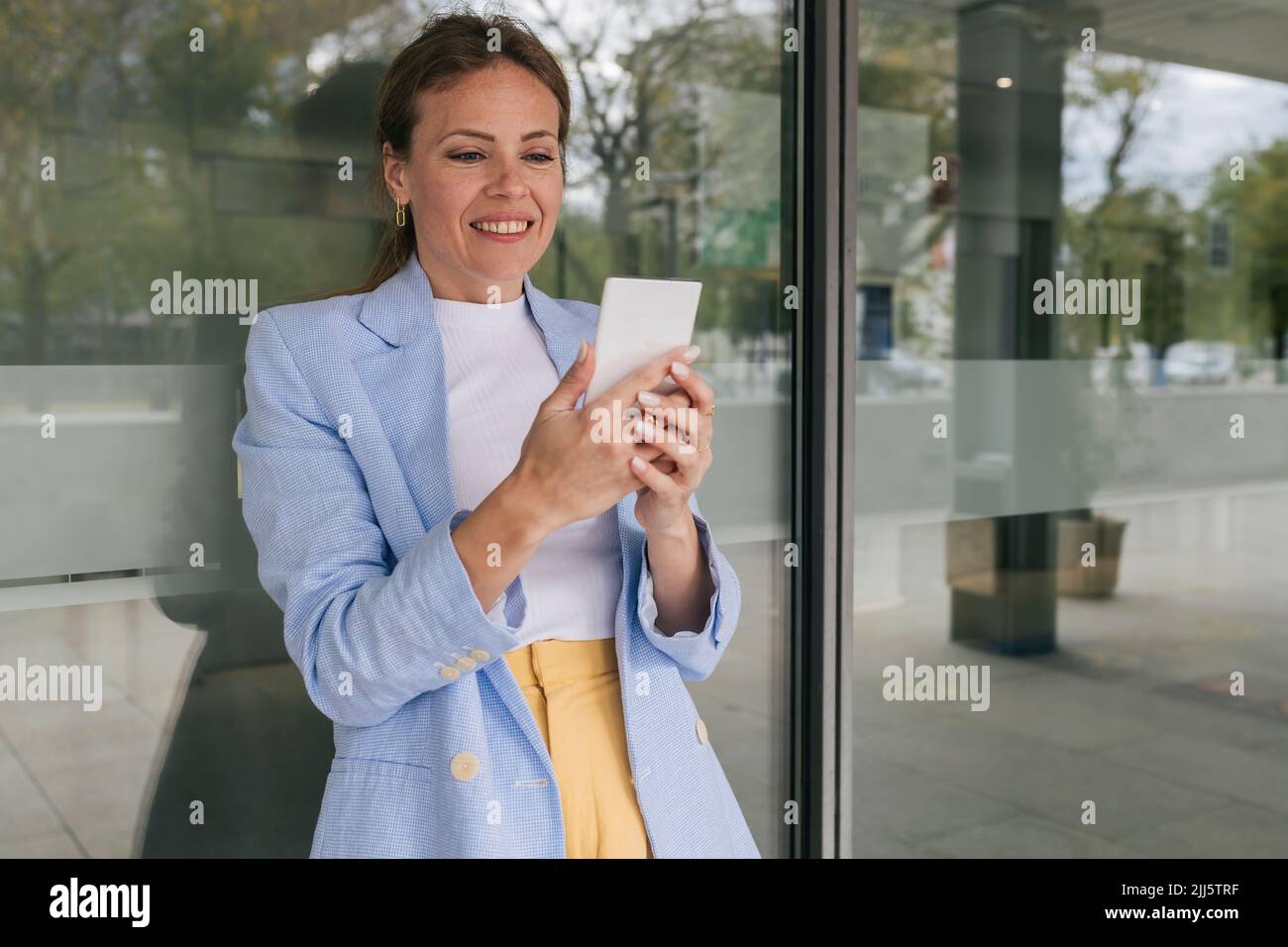 Smiling woman using phone leaning on glass window Stock Photo