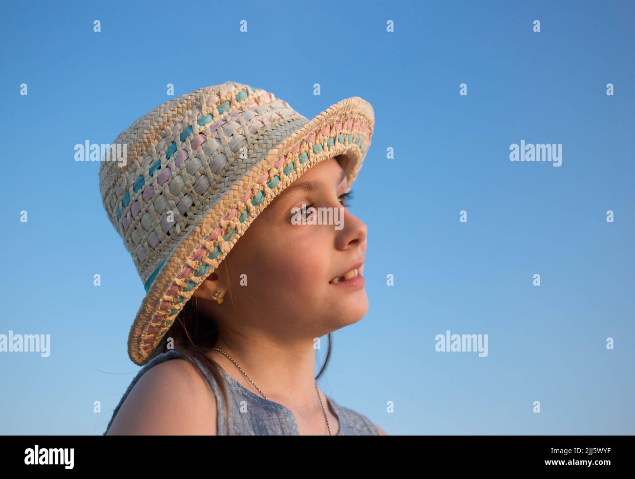 Portrait of a cute dreamy girl 6-7 years old in a straw hat against the blue sky. side view. profile. Romantic mood. childhood. vacation, summer time. Stock Photo