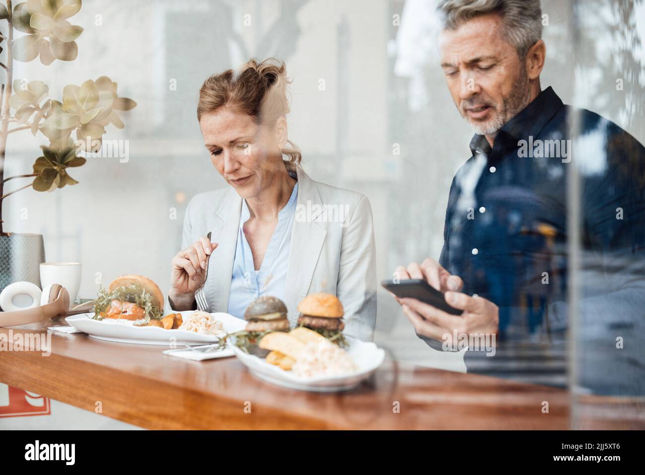 Businesswoman having lunch sitting by businessman using smart phone in cafe seen through glass Stock Photo