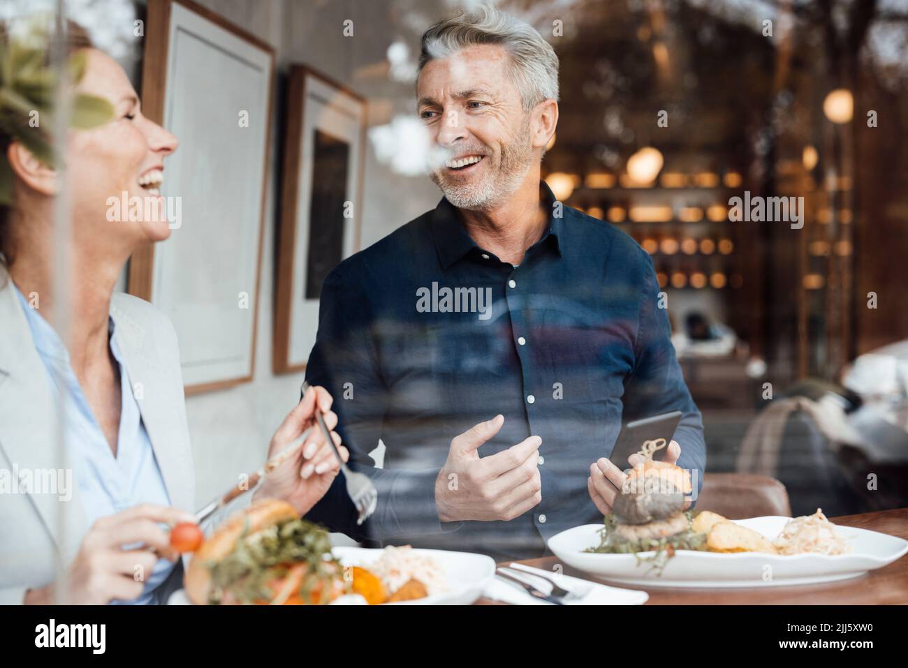 Happy businesswoman with businessman holding mobile phone having lunch in cafe seen through glass Stock Photo